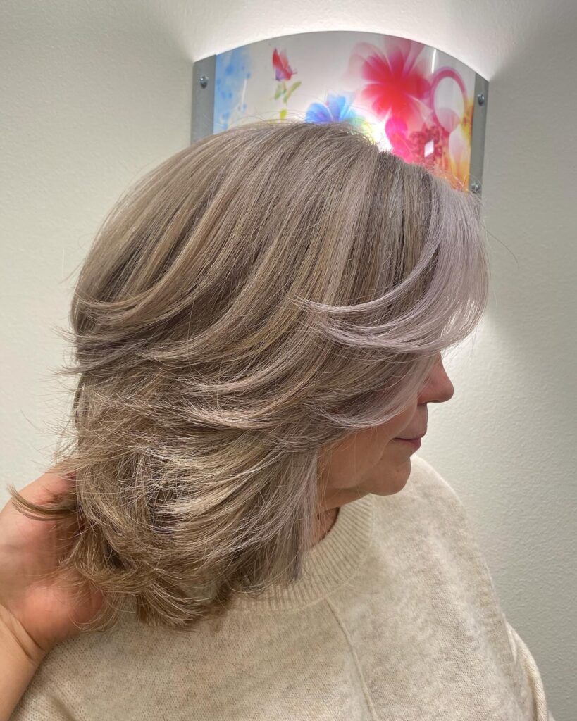 A woman is viewed from the side, showcasing a shoulder-length feathered shag haircut with ash blonde and silver layers.