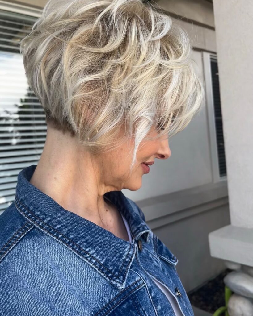 A side profile view of a woman over 60 wearing a denim jacket and sporting a voluminous platinum blonde textured wedge bob with soft waves.