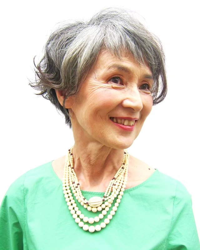 A smiling older woman with natural silver wavy hair and wispy bangs wearing a green top.