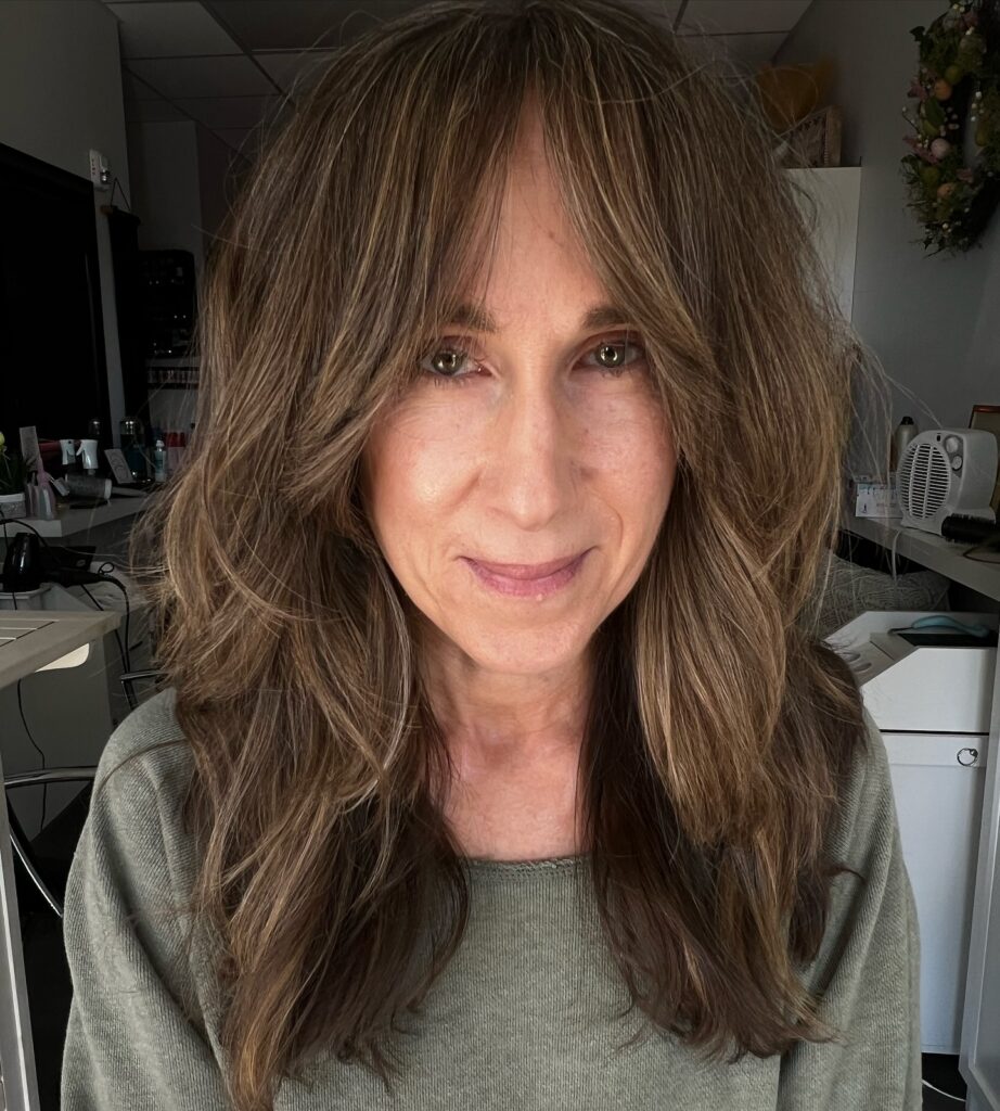 A front view of a smiling woman over 60 with long brunette layered hair featuring curtain bangs and subtle highlights.