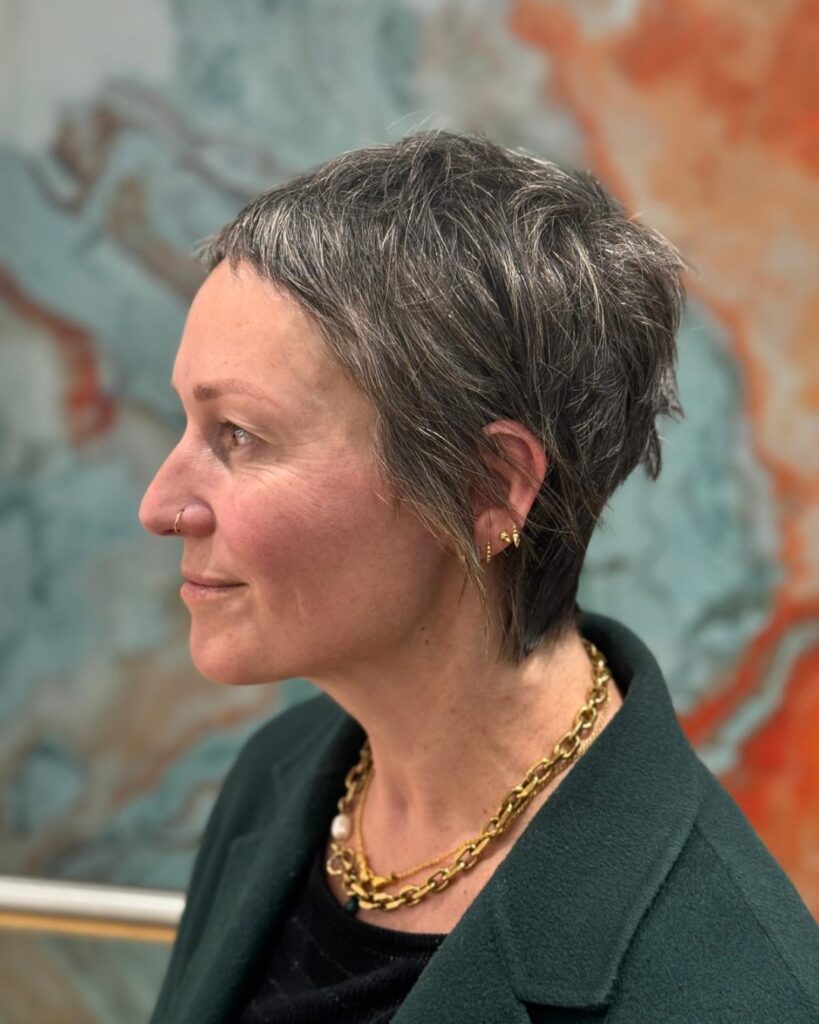 A side profile view of a woman over 60 with an edgy, short salt and pepper pixie haircut featuring textured baby bangs.
