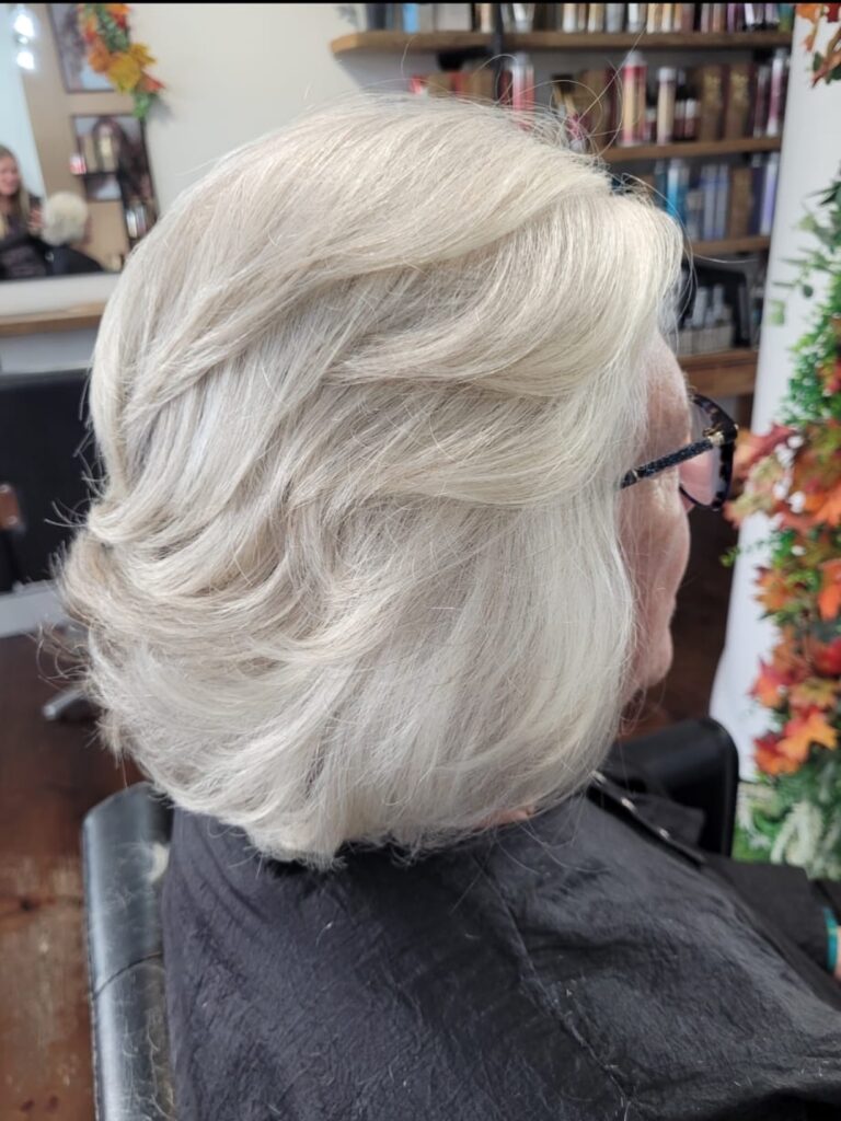 A side-angle view of a woman over 60 displaying a neck-length platinum-silver lob with voluminous feathered layers styled backwards.