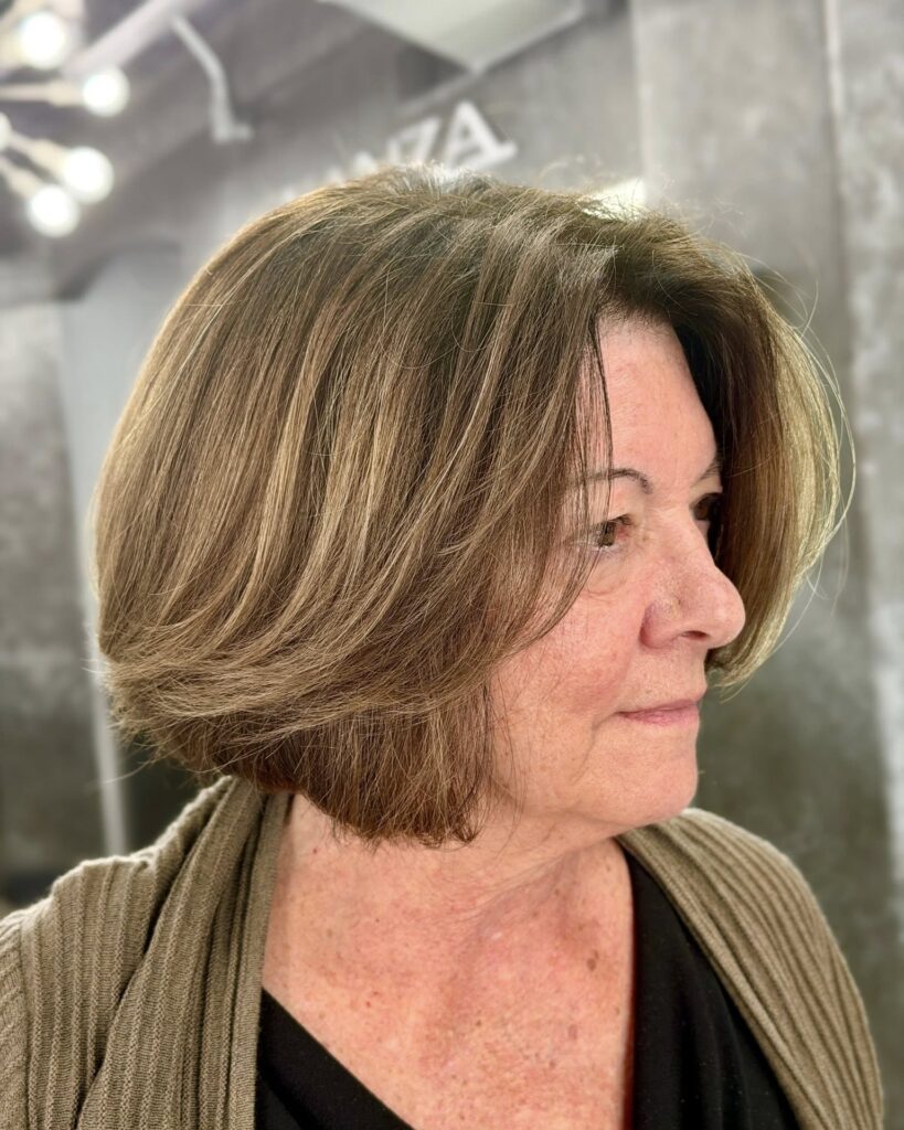 A side profile view of a woman over 60 with a chin-length brunette bob featuring warm caramel highlights and face-framing layers.
