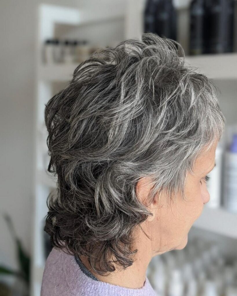 A side profile view of a woman over 60 showcasing a textured, salt-and-pepper shaggy mullet haircut with natural waves.