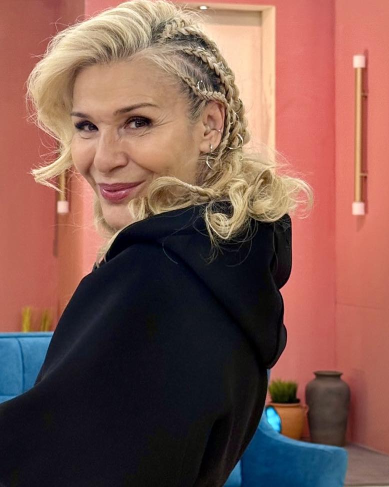 A smiling woman over 60 looking over her shoulder, showcasing a half-braided hairstyle with long blonde curls and decorative hair rings.