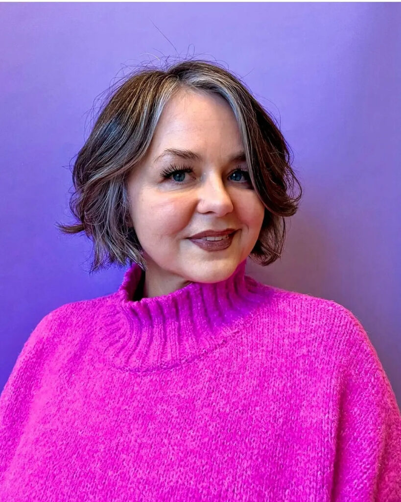 A front view of a smiling woman over 60 wearing a hot pink sweater and showcasing a wavy brunette bob with a bright blonde money piece highlight.