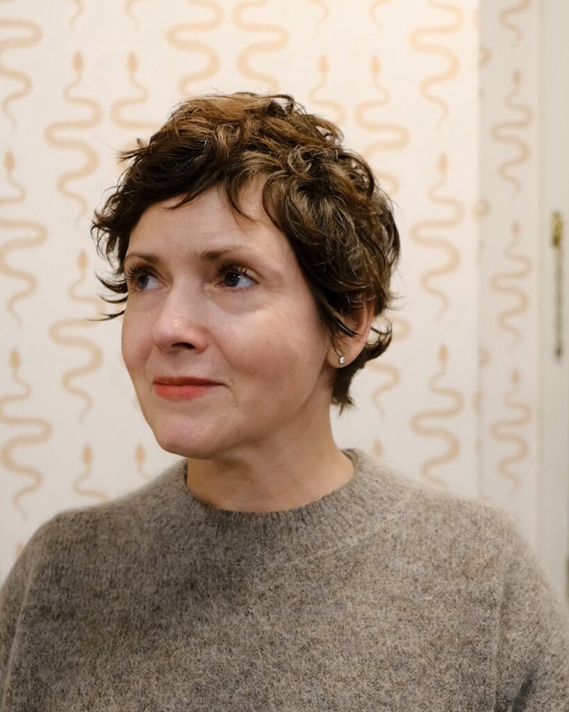 A three-quarter profile view of a smiling woman over 60 with a short, messy, curly brunette pixie haircut.
