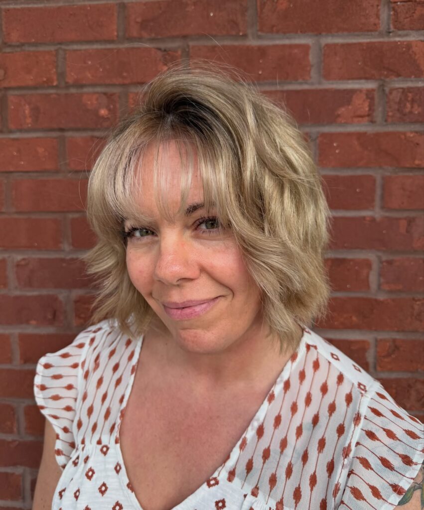 A smiling woman with a sandy blonde textured lob haircut featuring loose beachy waves and wispy bangs.