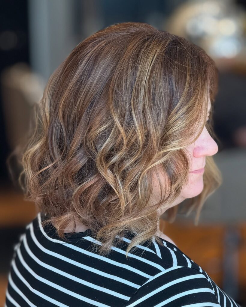 A woman showing a side profile of a neck length caramel blonde wavy lob with sun kissed highlights.