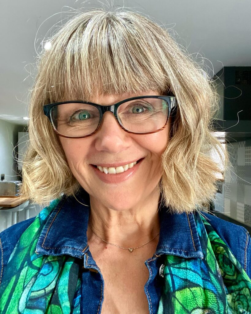 A smiling woman with a chin length honey blonde wavy bob and full straight bangs.