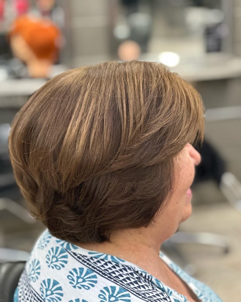 A side profile view of a woman showcasing a honey chestnut brown pixie bob haircut with feathered layers swept back.