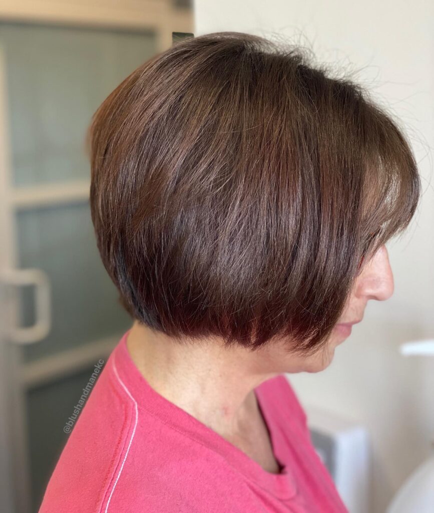 A side profile view of a woman showcasing a sleek, chin-length auburn brown French bob haircut with a smooth straight texture.