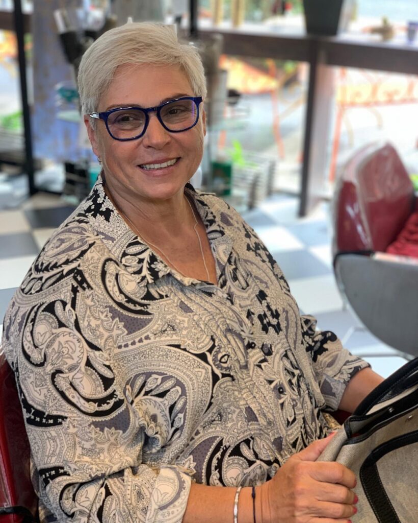 A smiling woman over 60 with a short silver side-swept pixie haircut and blue glasses.
