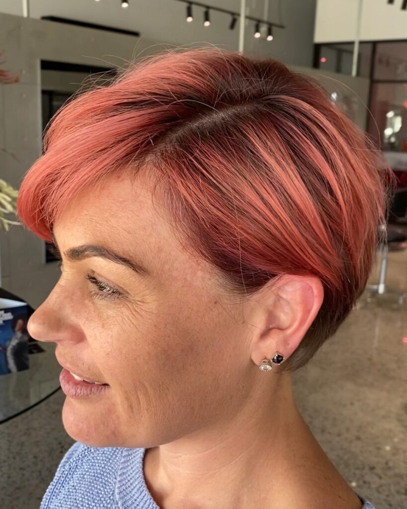 A side profile of a woman with a sleek, side-parted pixie cut dyed a vibrant coral pink color.