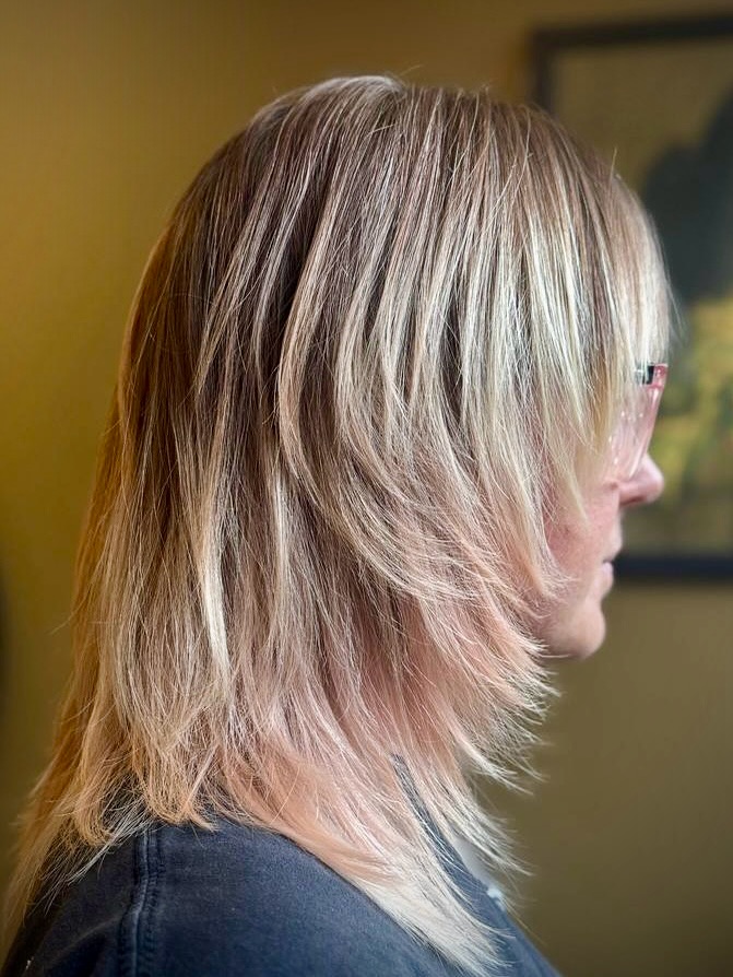 A side profile view of a woman with straight shoulder-length blonde hair featuring heavily layered, feathered face-framing pieces and subtle pink undertones at the tips.