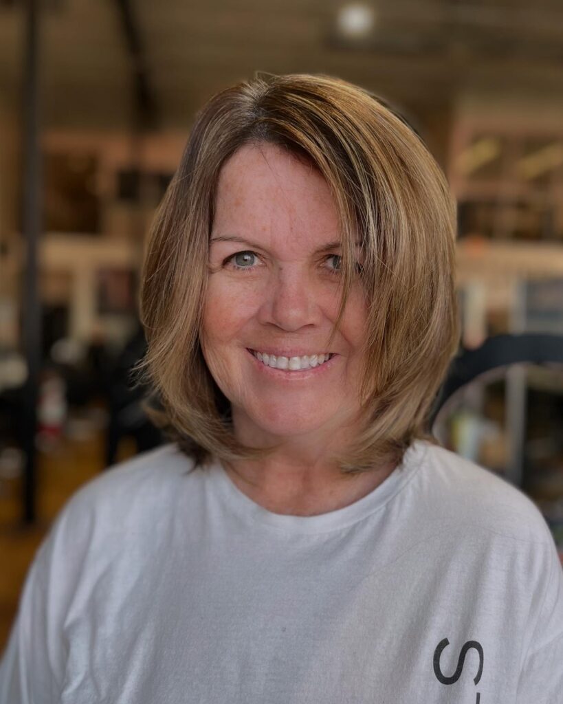 A smiling woman with a shoulder-length layered long bob hairstyle featuring warm golden blonde highlights and face-framing layers.