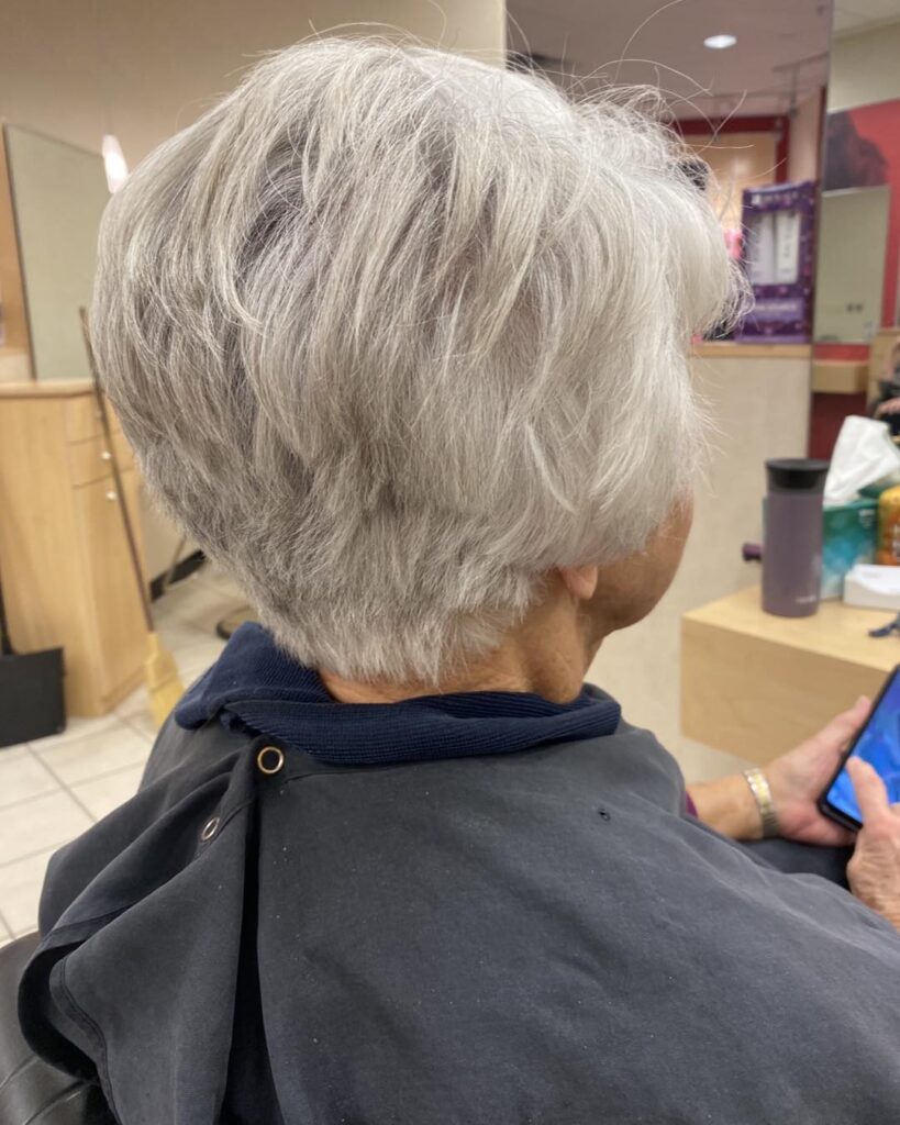 A back view of a woman with a short feathered silver pixie bob haircut featuring piecey layers and a tapered neckline.