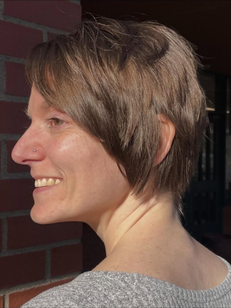 A side profile view of a smiling woman with a short ash brunette pixie bob haircut featuring wispy layers and a textured side fringe.