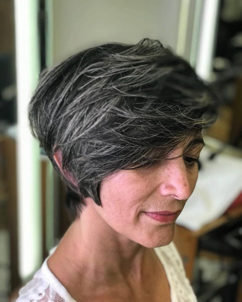 A side profile view of a woman with a texturized charcoal gray pixie haircut featuring bright silver highlights and a long side-swept fringe.
