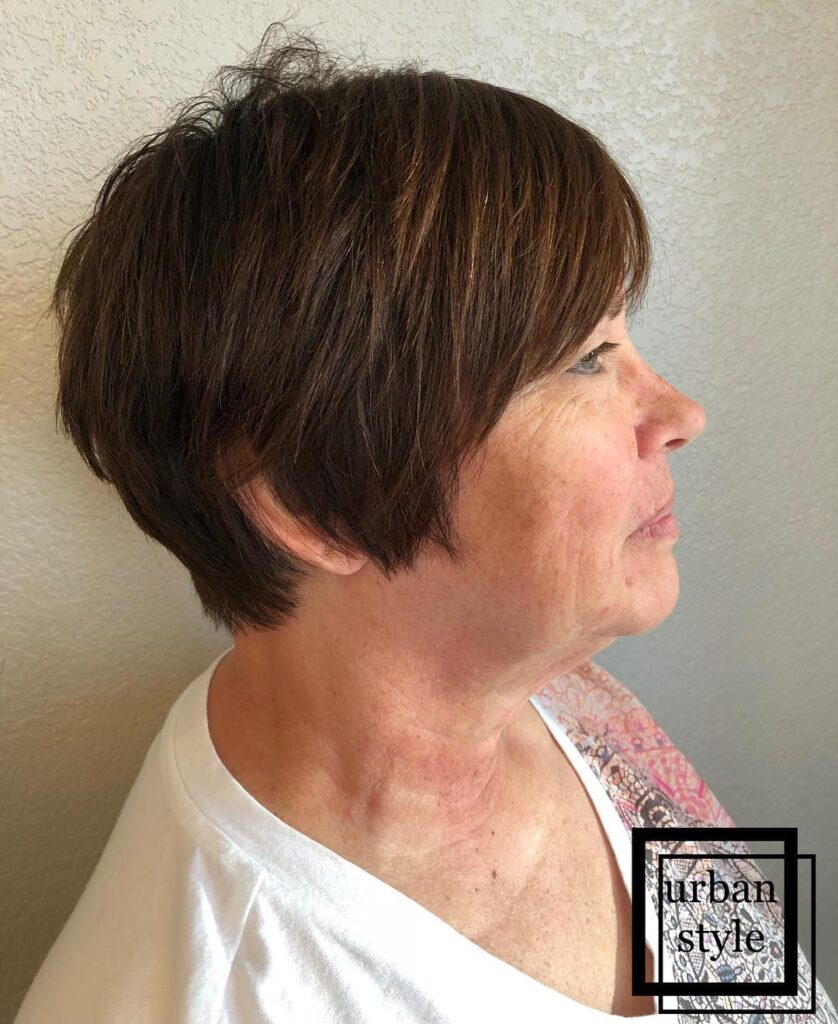 A side profile view of a woman with a short tapered brunette pixie haircut featuring soft side-swept bangs and subtle caramel highlights.