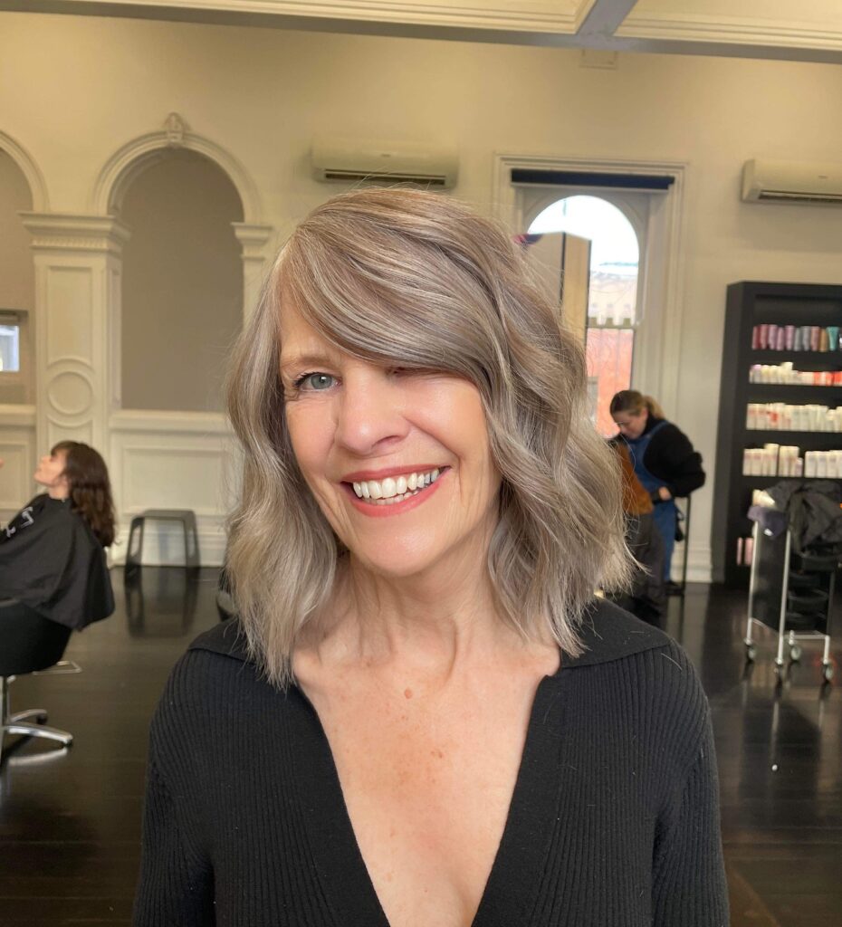A smiling woman with a shoulder-length wavy mushroom-grey lob and a deep side-swept fringe.