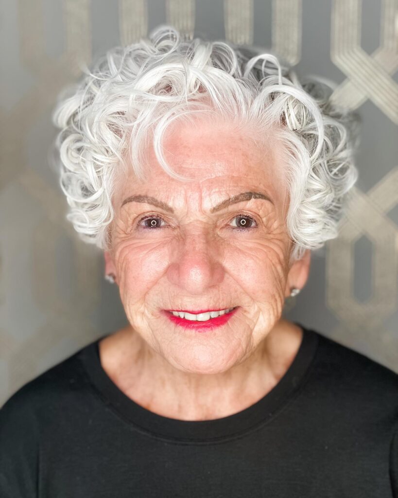 A close-up portrait of an older woman smiling with voluminous short white curly hair.