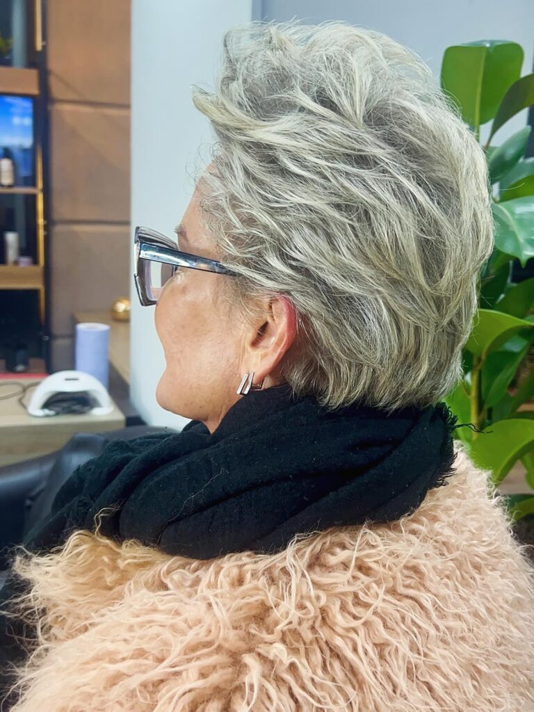 A side view of a woman wearing glasses with a voluminous short ash-grey hairstyle featuring swept-back layers.