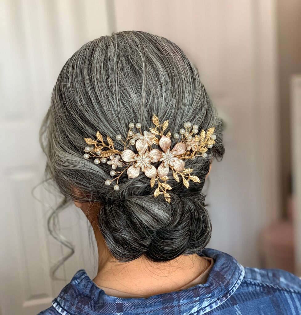 A back view of a woman with salt-and-pepper hair styled in a low chignon adorned with a gold floral hairpiece.