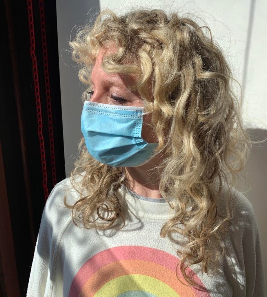 A woman in profile view wearing a face mask and a rainbow sweatshirt displaying her medium-length honey blonde curly shag haircut with curly bangs.
