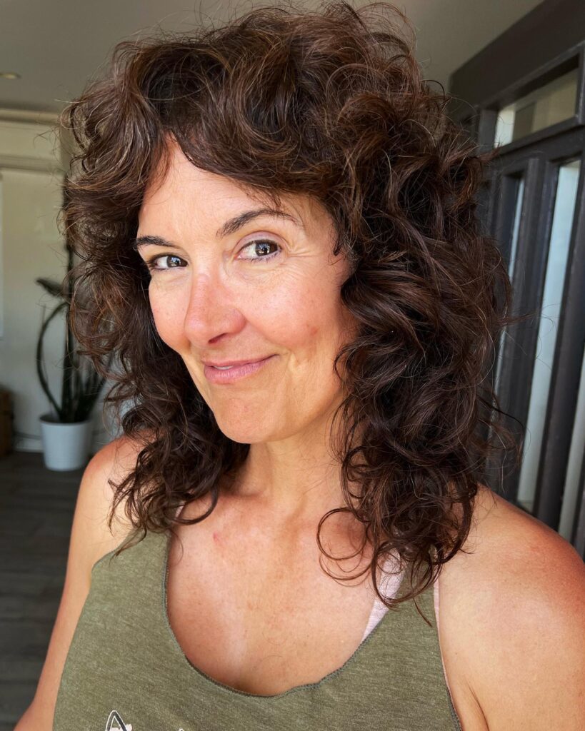 A smiling woman with shoulder-length shaggy curly brown hair looking to the side.