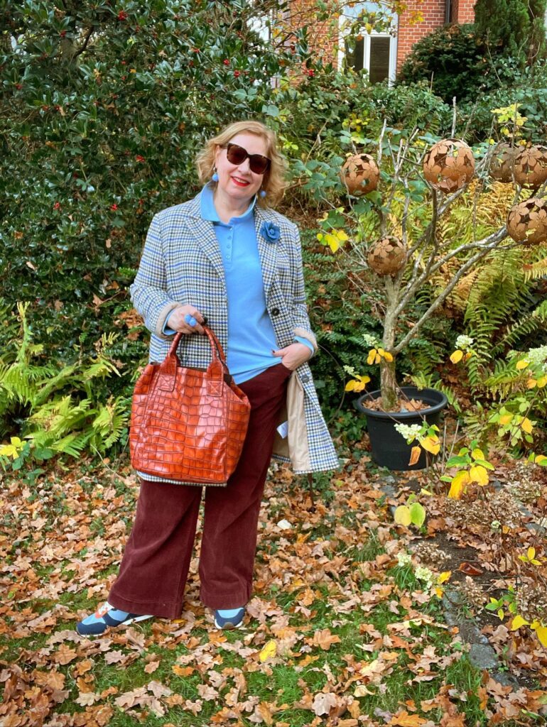 A woman with shoulder-length honey blonde curly hair wearing sunglasses and a patterned coat in a garden.