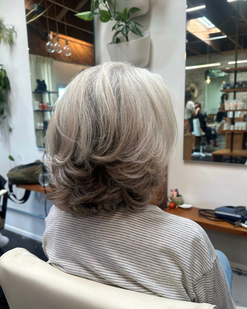 A back view of a woman sitting in a salon chair showcasing a shoulder-length feathered silver and blonde haircut with stacked layers.