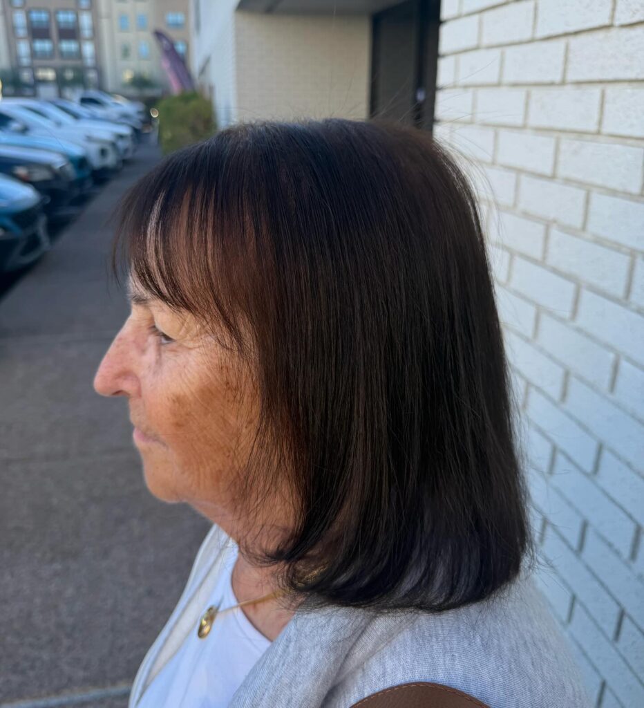 A side profile view of a woman outdoors showcasing a straight shoulder length dark brunette bob with light wispy bangs.