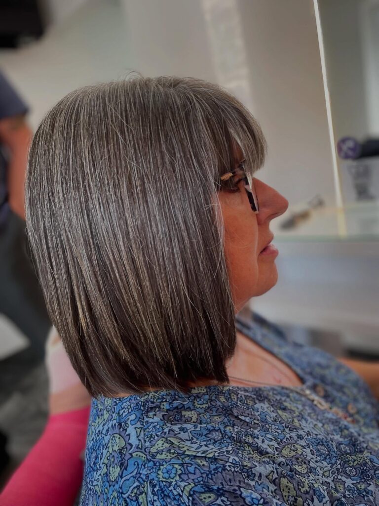 A side view of a woman with a sleek shoulder-length salt and pepper haircut and full straight bangs.