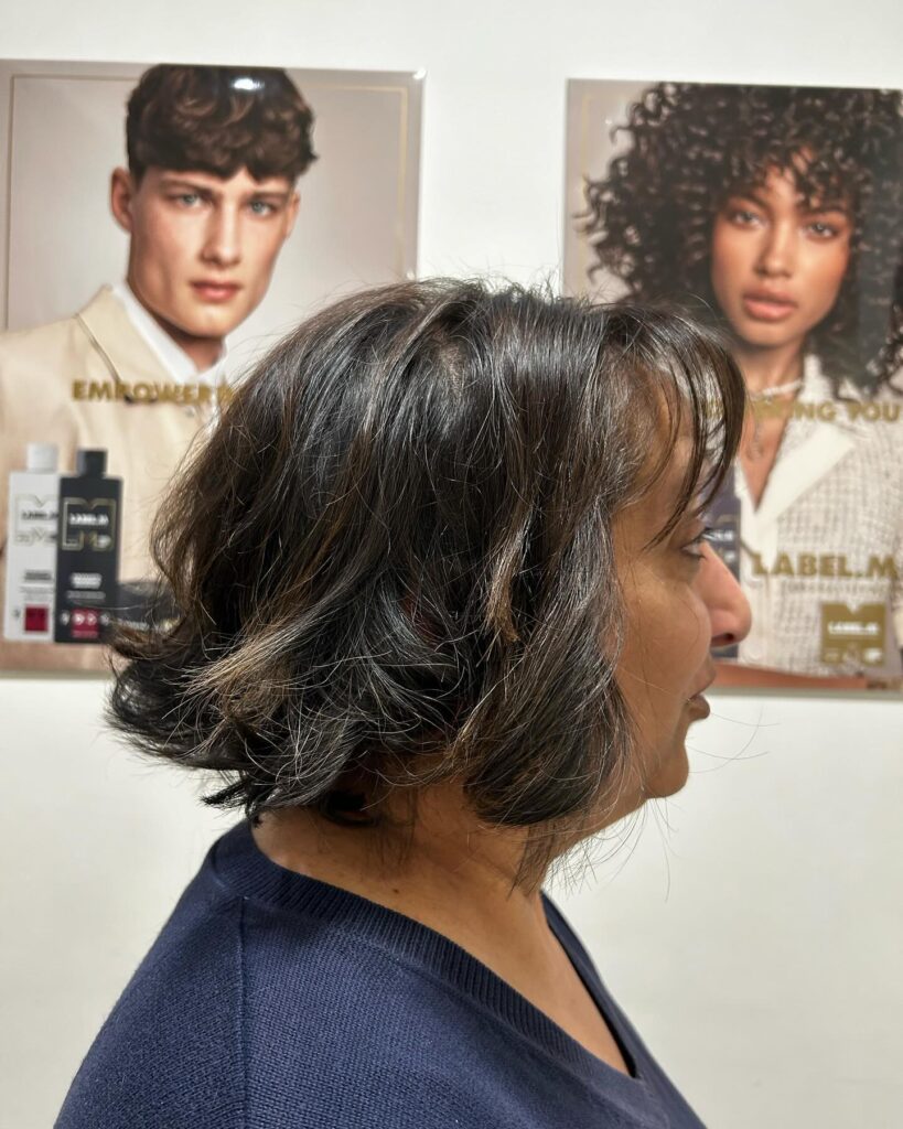 A woman with fine, dark brunette hair showcases a textured chin-length inverted bob with light bangs and subtle highlights from a side view.