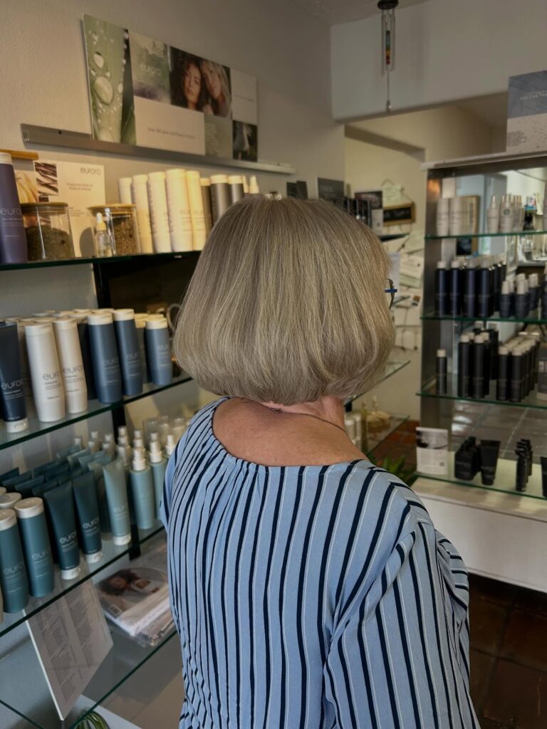An older woman viewed from behind displays a classic neck-length ash blonde bob with smooth, under-tucked ends.