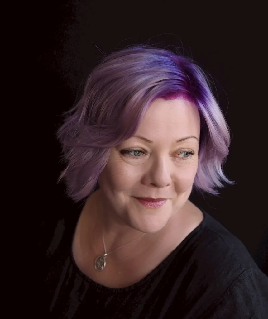 A woman with green eyes smiles while showcasing a textured chin-length lavender and violet bob with soft waves and dark magenta roots.