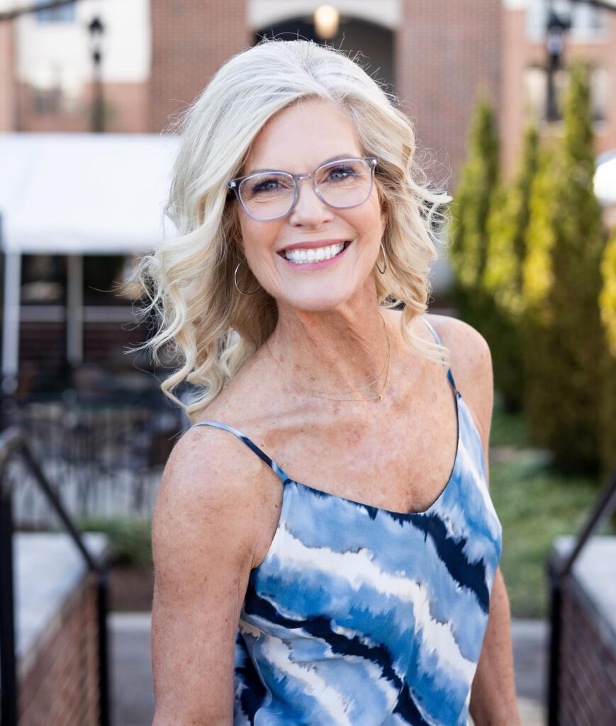 A smiling woman over 60 wearing glasses showcases a voluminous shoulder-length platinum blonde hairstyle with elegant waves and a deep side part.