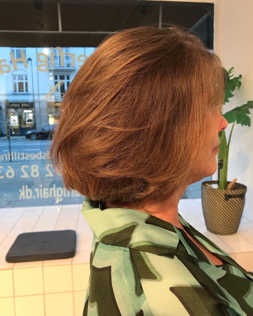 A woman from a side profile view displays a rounded neck-length auburn bob haircut with soft layers and warm golden highlights.