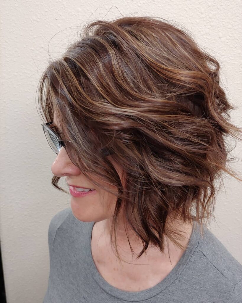A woman from a side profile view displays a textured chin-length brunette bob with choppy layers and warm caramel highlights.