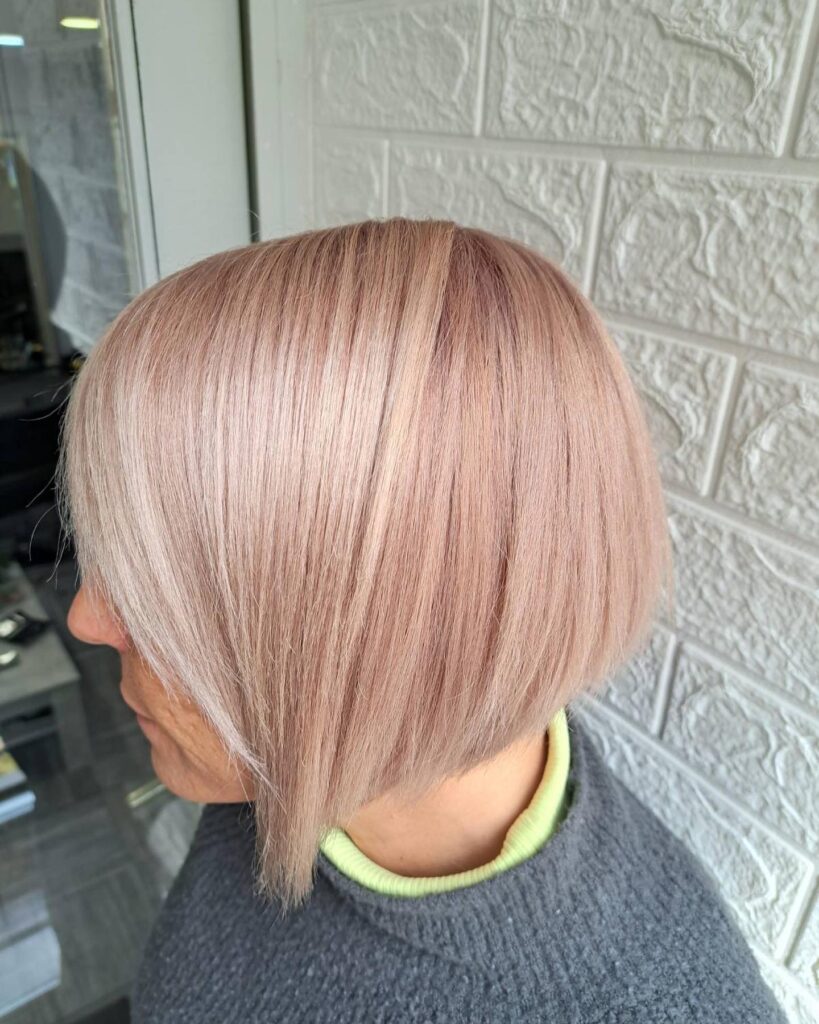 A woman from a side profile view displays a sleek, straight asymmetrical jaw-length bob haircut in a pastel rose gold color.