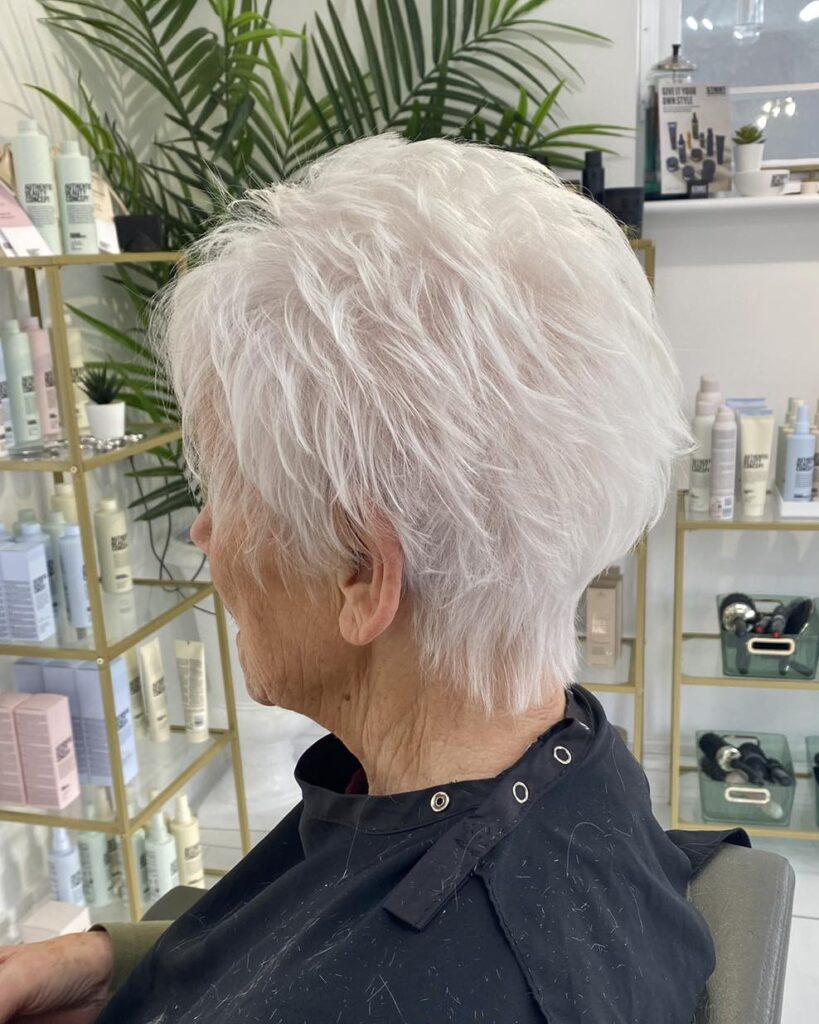 A woman viewed from the side showcasing a short, textured white pixie cut with feathery layers at a salon.