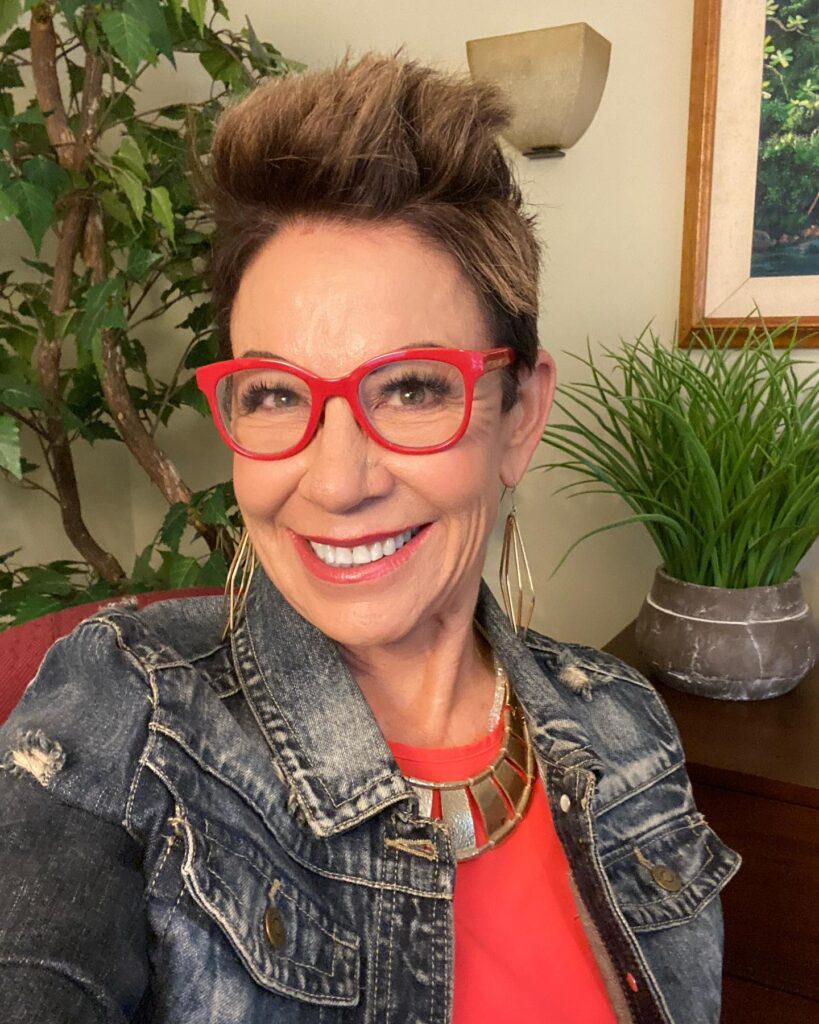A smiling woman over 60 with a short, spiky brunette pixie cut styled high on top wearing red glasses and a denim jacket.