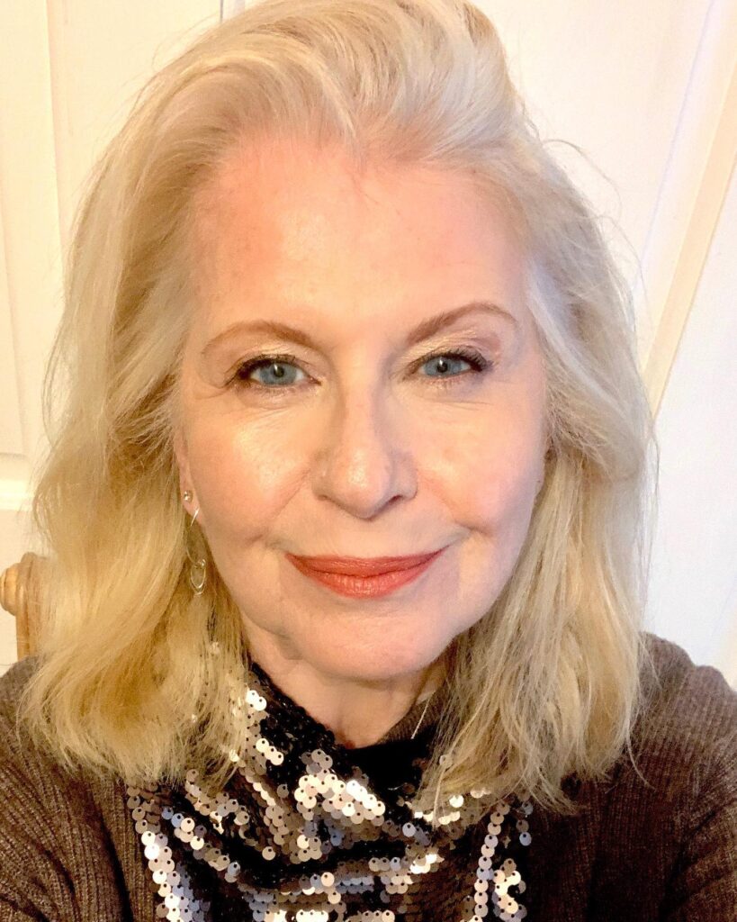 A smiling woman over 60 with a shoulder length golden platinum lob haircut styled swept back with volume at the roots.