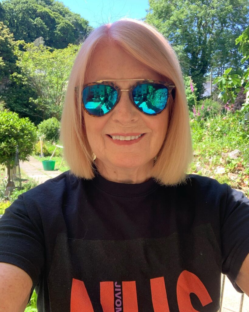 A smiling woman over 60 with a straight, neck length peach blonde bob haircut wearing sunglasses outdoors.