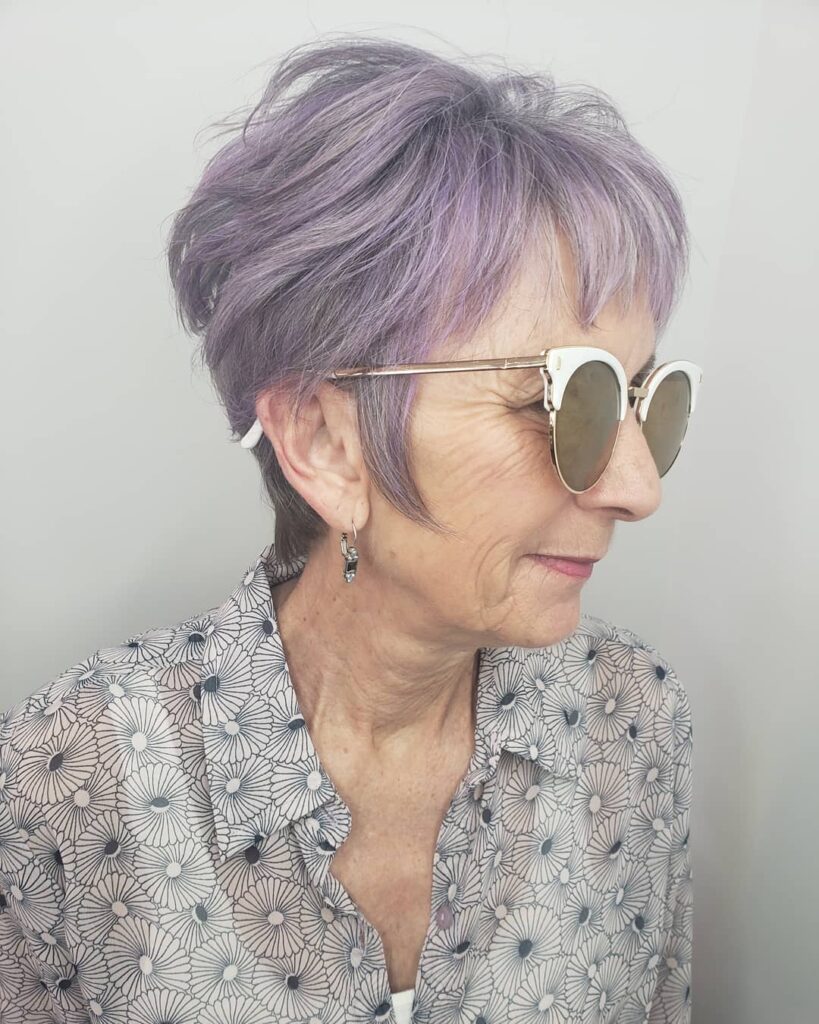 A side profile photograph of a woman with a choppy layered pixie haircut in a dusty lilac color wearing stylish white sunglasses.