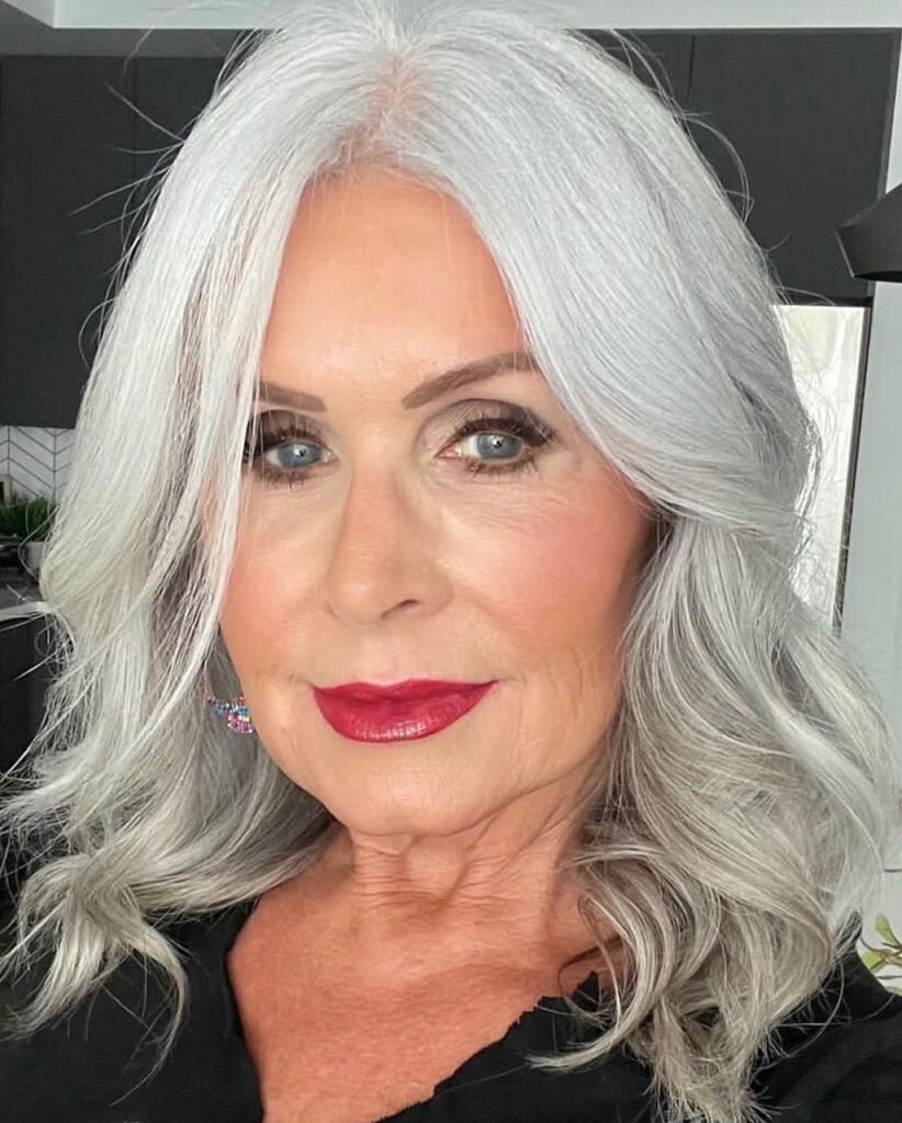 A woman over 60 with mid length silver hair styled in a bouncy, voluminous blowout with face framing layers.