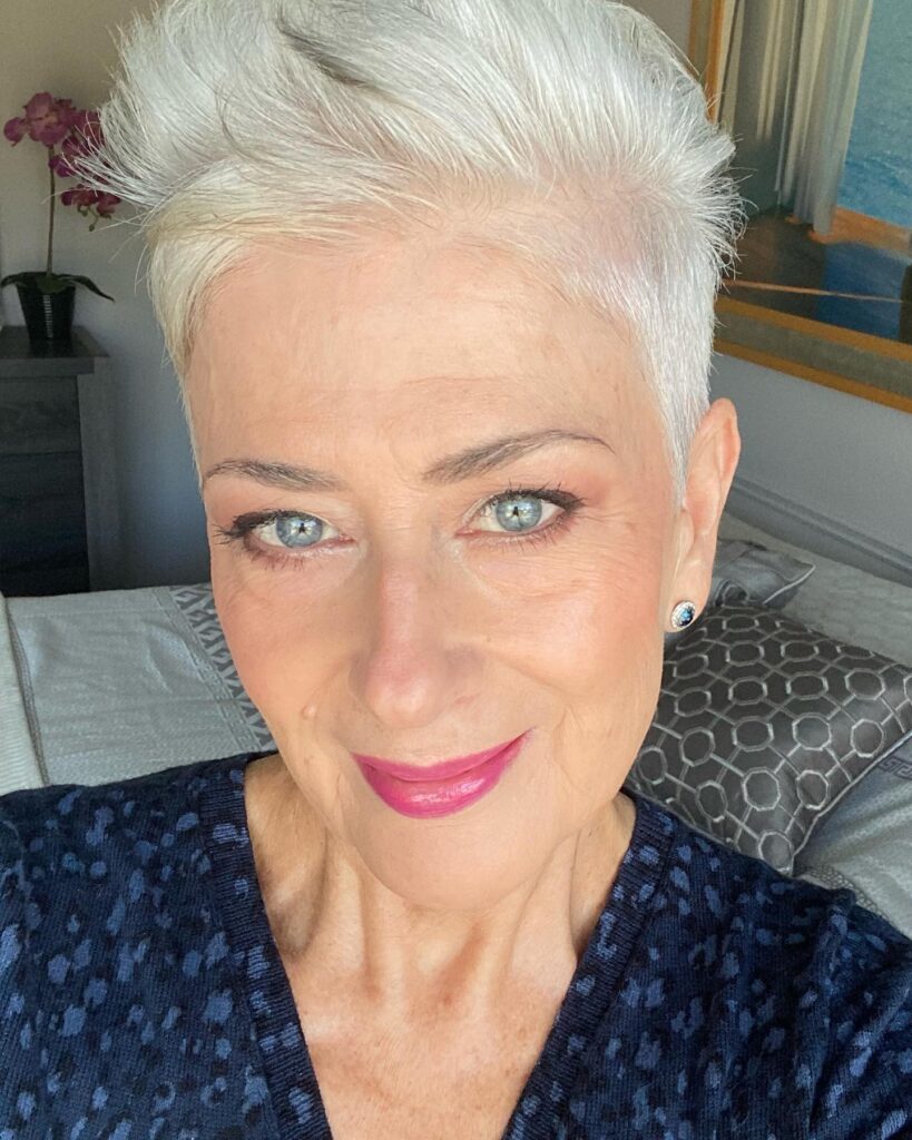A close up photograph of a smiling woman over 60 showcasing a short, high volume platinum pixie cut with an upward styled pompadour.
