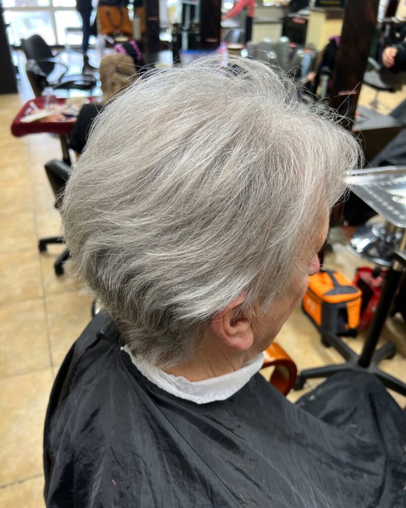 An over-the-shoulder photograph of a woman with a short, tapered silver grey haircut styled in soft, feathery layers.