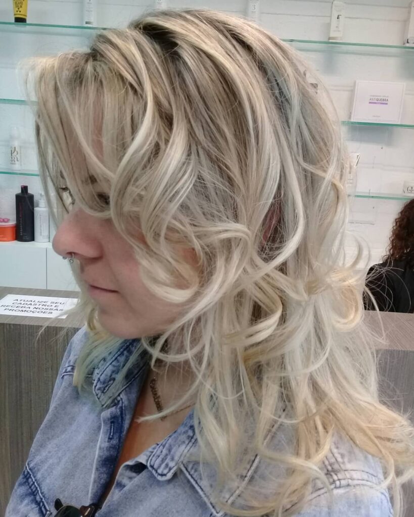 A woman with mid-length layered wavy pearl blonde hair featuring voluminous curls and face-framing pieces.